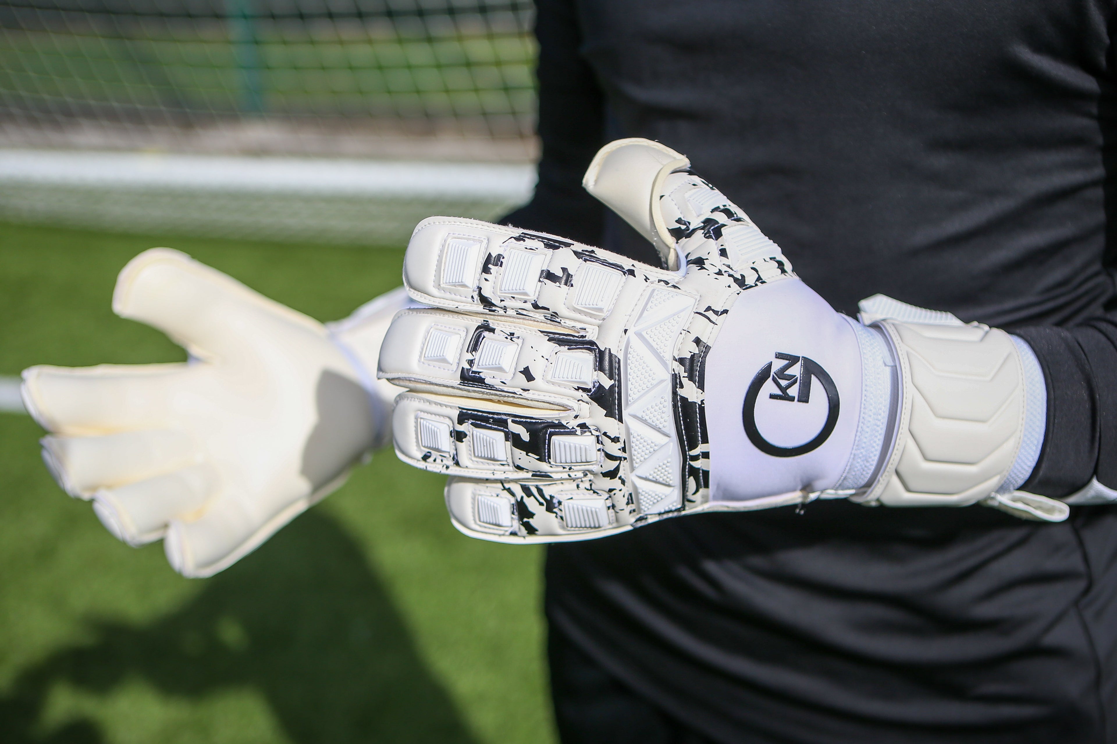 Close-up of a white GKN football glove with black camouflage details on the fingers. The GKN logo is on the wrist, and the glove has padded support for grip and flexibility, shown against the background of a football goal."