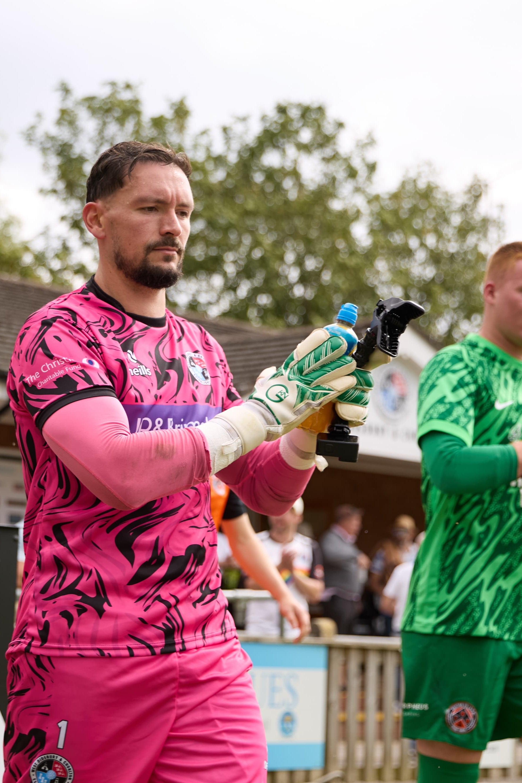 Soccer goalkeeper in pink kit with gloves, surrounded by other players and spectators outdoors.