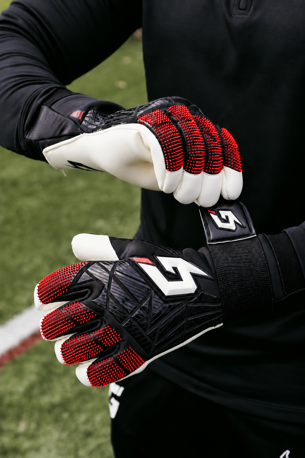 Close-up of a person wearing black, white, and red soccer goalkeeper gloves, adjusting the wrist strap of one glove on an outdoor grass field.